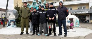 Eine Gruppe uniformierter Feuerwehrleute und Kinder steht vor einem Gebäude mit einer schneebedeckten Iglu-Skulptur. Die Feuerwehrleute tragen Mützen und halten eine Laterne. Die Kinder sind in Winterkleidung gekleidet.