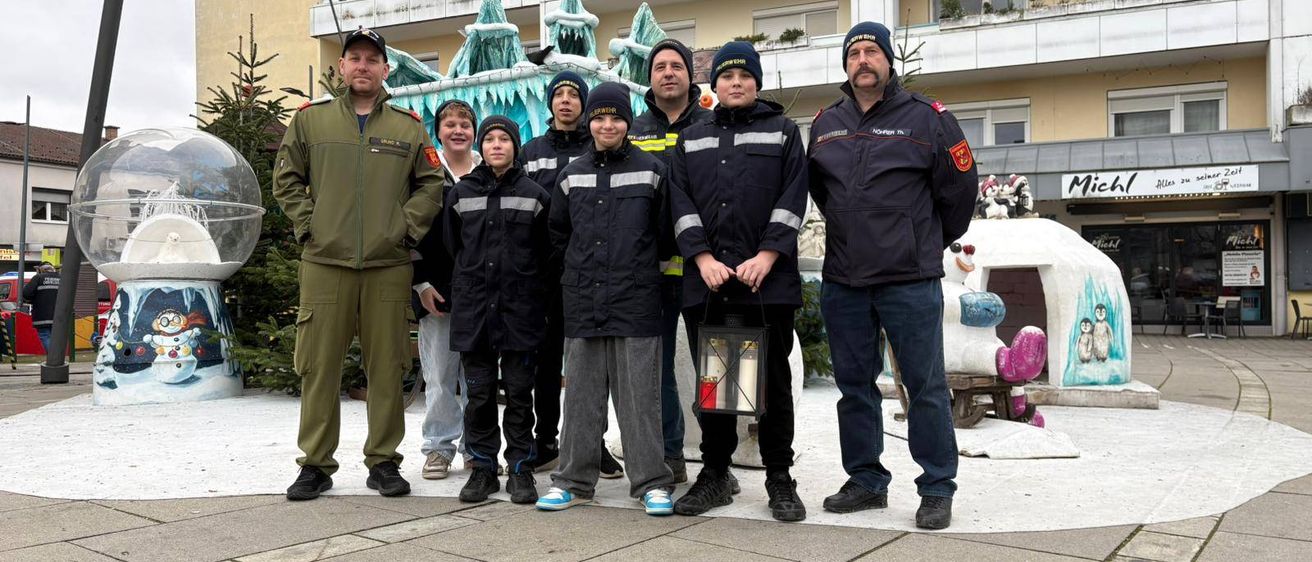 Eine Gruppe uniformierter Feuerwehrleute und Kinder steht vor einem Gebäude mit einer schneebedeckten Iglu-Skulptur. Die Feuerwehrleute tragen Mützen und halten eine Laterne. Die Kinder sind in Winterkleidung gekleidet.