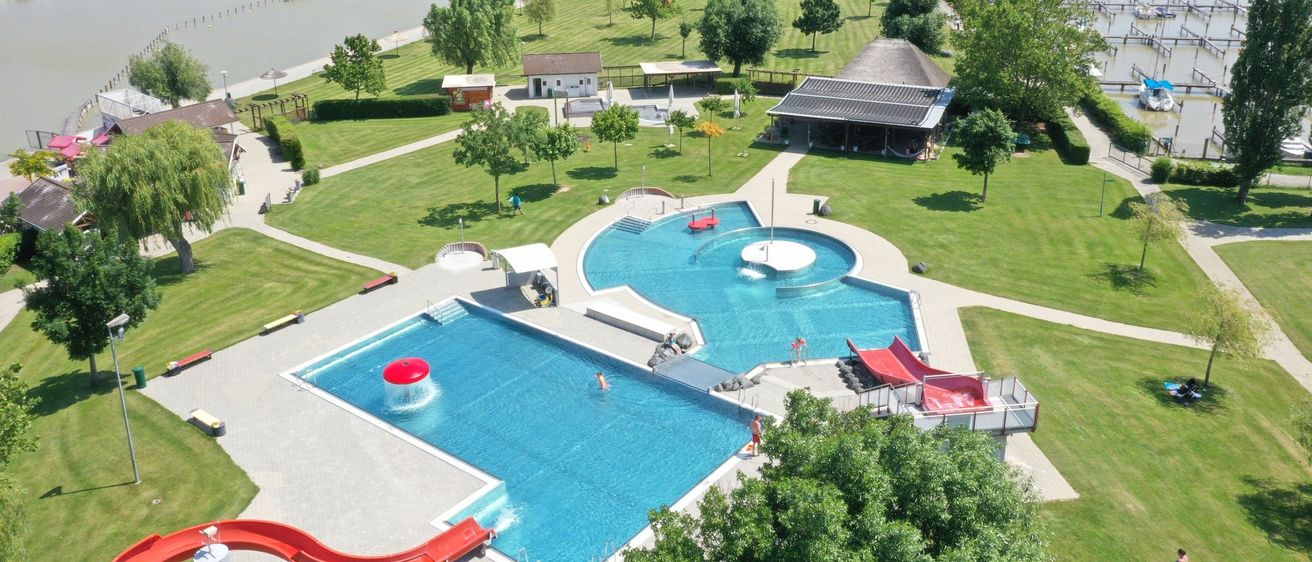 Luftaufnahme eines großen Freizeitparks mit Schwimmbad, Wasserrutschen und Strandbereich. Bäume und ein Gewässer im Hintergrund.