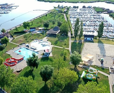 Luftaufnahme eines Erholungsgebiets mit Schwimmbad, Wasserrutsche, Spielplatz und vielen gedockten Booten in einer Marina.