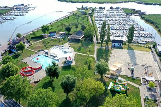 Luftaufnahme eines Erholungsgebiets mit Schwimmbad, Wasserrutsche, Spielplatz und vielen gedockten Booten in einer Marina.