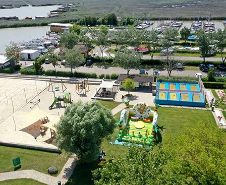 Luftaufnahme eines Spielplatzes mit Volleyballfeld, Spielgeräten und einem Sprungburg. Im Hintergrund ein Hafen mit angedockten Booten.