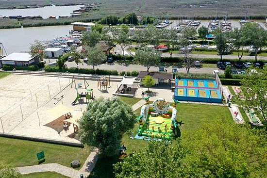 Luftaufnahme eines Spielplatzes mit Volleyballfeld, Spielgeräten und einem Sprungburg. Im Hintergrund ein Hafen mit angedockten Booten.