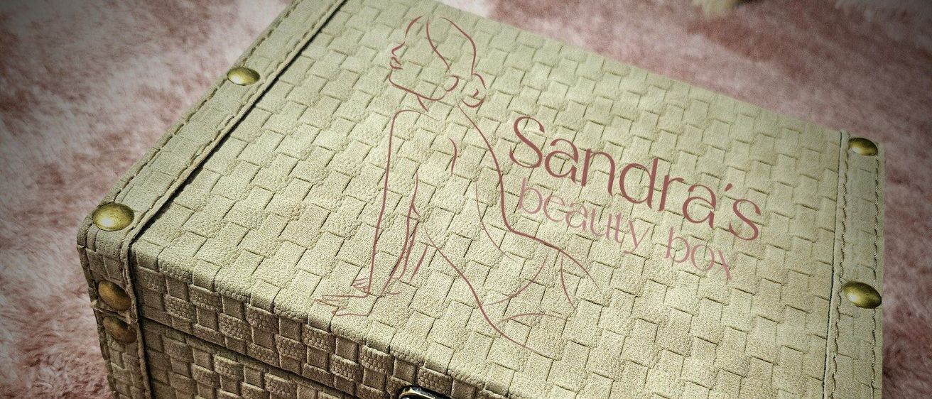 Eine beige Schönheitsbox mit dem Namen 'Sandra's Beauty Box' mit einem Flechtmuster und einem Schloss oben liegt auf einem rosa Teppich.