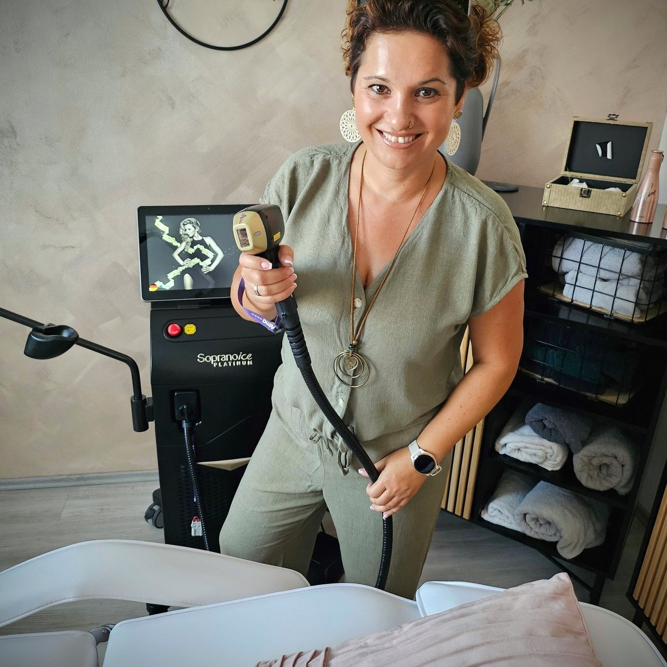 Eine Frau in einem grünen Overall steht in einem Raum mit einem Lasergerät und lächelt. Sie hält das Gerät mit einem Bildschirm, auf dem ein Bild einer Frau angezeigt wird. Ein Regal hält Handtücher und ein hölzerner Schrank hat eine Schachtel obenauf.