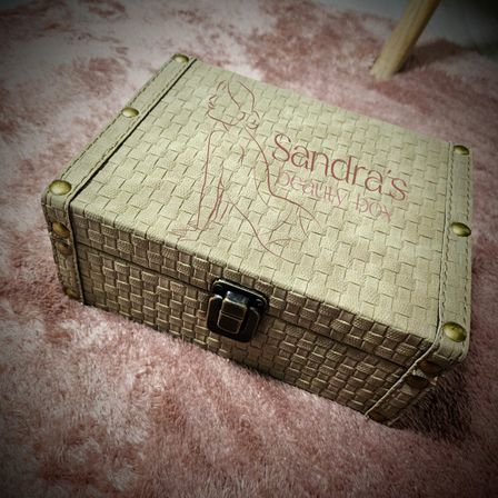 Eine beige Lederbox mit der Aufschrift 'Sandra's Beauty Box' liegt auf einem rosa Teppich. Die Box hat ein gewebtes Muster und Messingfarbene Beschläge.