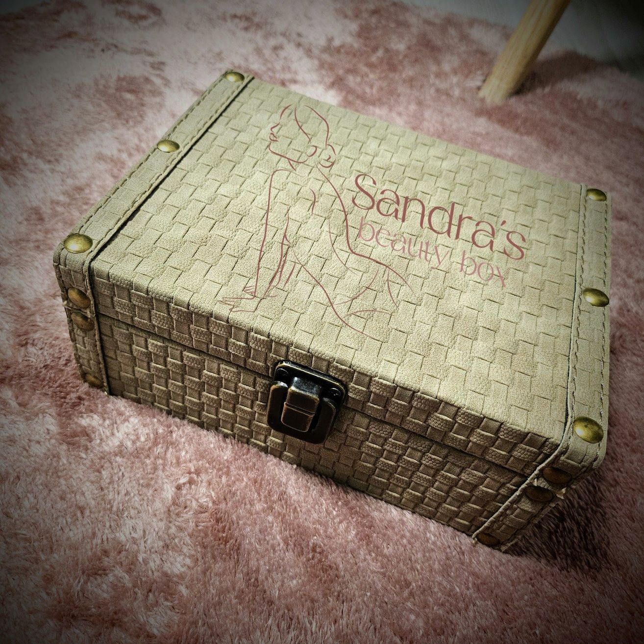 Eine beige Lederbox mit der Aufschrift 'Sandra's Beauty Box' liegt auf einem rosa Teppich. Die Box hat ein gewebtes Muster und Messingfarbene Beschläge.