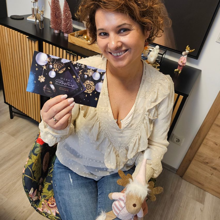 Eine Frau hält eine Weihnachtsgeschenkbox und einen Plüschrentier, lächelt und sitzt auf einem Stuhl in einem dekorierten Raum.