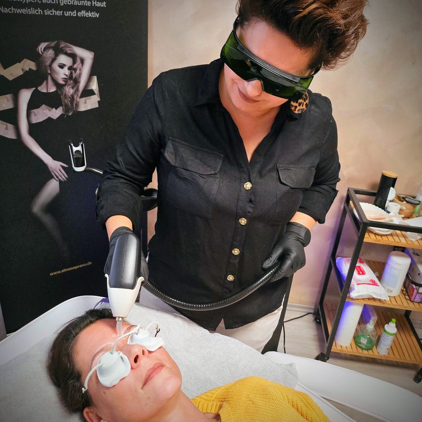 Eine Frau in einem Salon, die eine Brille trägt, verabreicht einer liegenden Kundin mit Schutzbrille eine Behandlung. Im Hintergrund ist ein Poster einer Frau in einem schwarzen Kleid zu sehen.