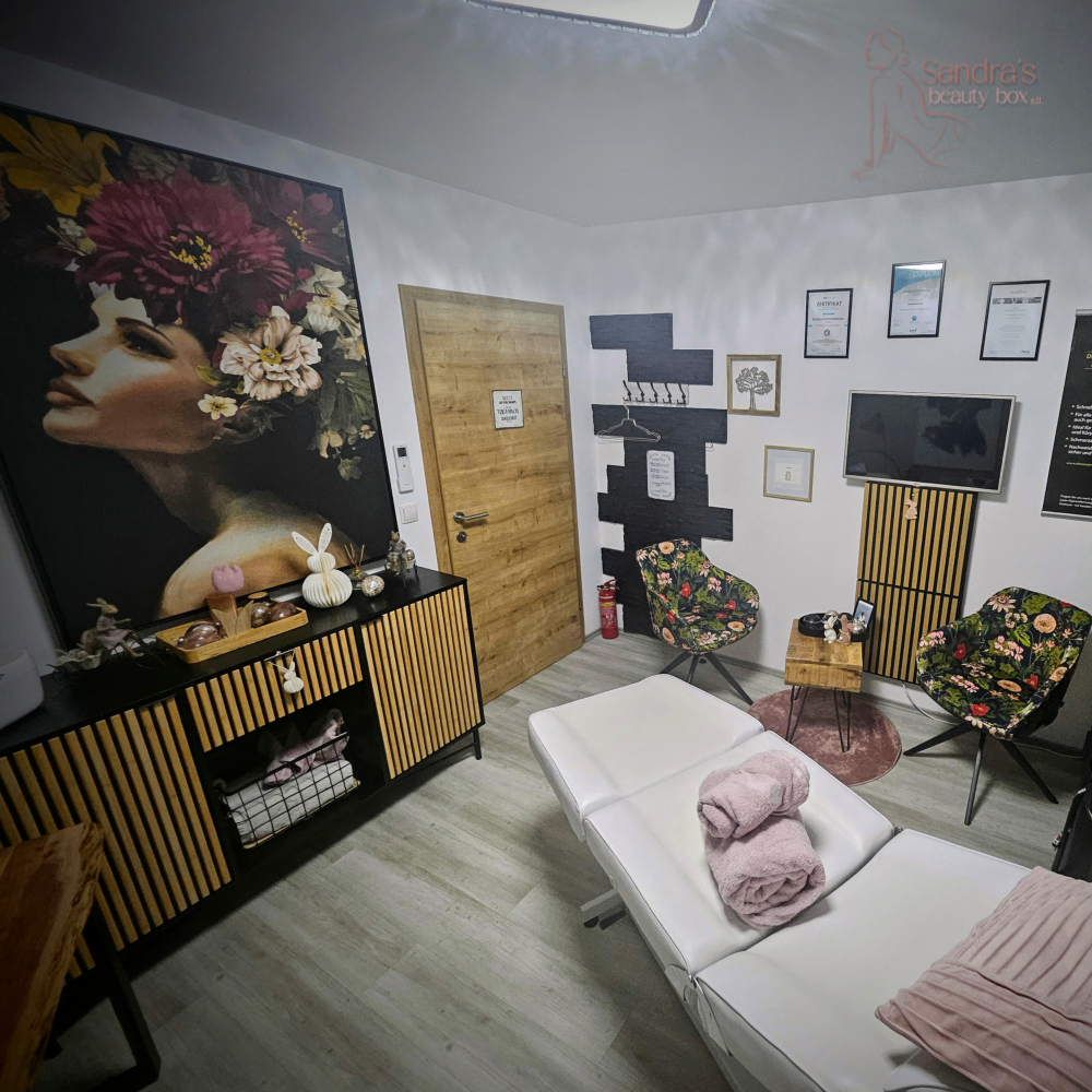 Innenansicht eines Schönheitssalons mit einem großen Gemälde einer blumengekrönten Frau, einer Holztür, Blumenstühlen, einem Schreibtisch mit Handtüchern, einem Holzschrank und einem an der Wand montierten Fernseher.