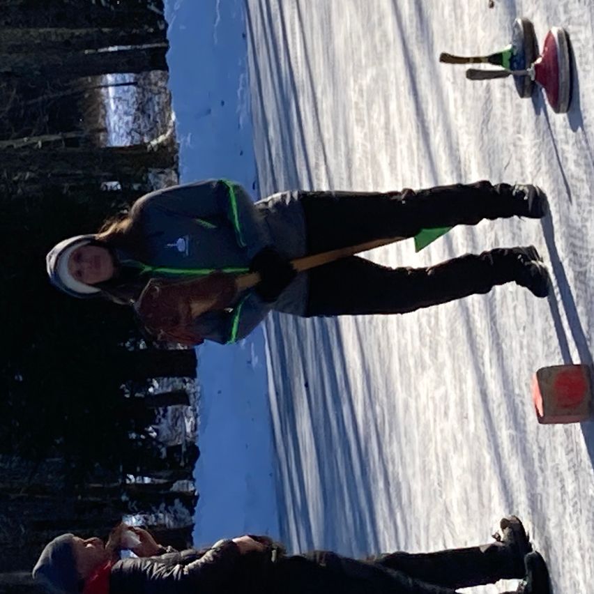Zwei Personen stehen auf einer Eisbahn und tragen Winterkleidung. Eine Person hält einen Besen. Es gibt drei Curling-Steine auf dem Eis.