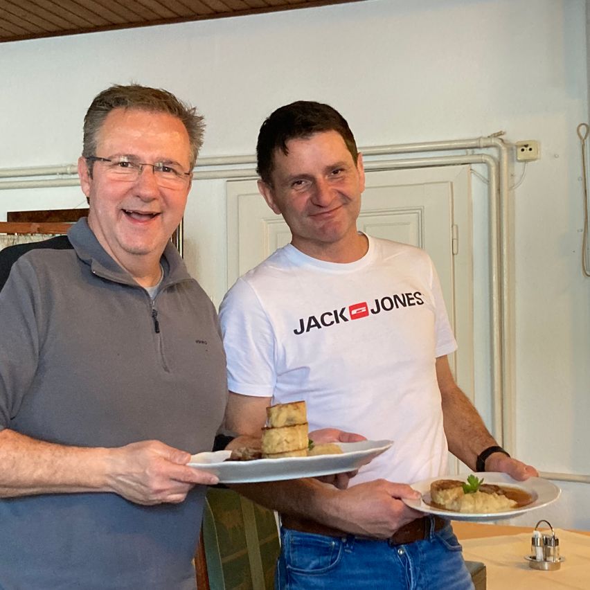 Zwei lächelnde Männer, einer in einem grauen Shirt und der andere in einem weißen Jack Jones T-Shirt, jeder hält einen Teller mit Essen vor einer weißen Wand.