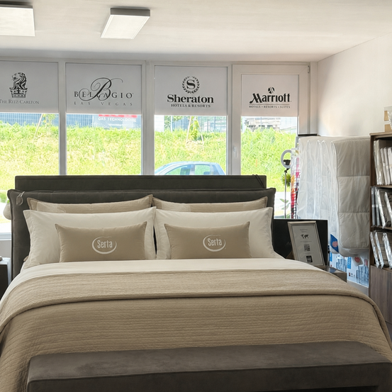 Schlafzimmer mit Serta-Kissen, einer beigefarbenen Decke und einem Fenster mit Logos für The Ritz-Carlton, Bellagio, Sheraton und Marriott.