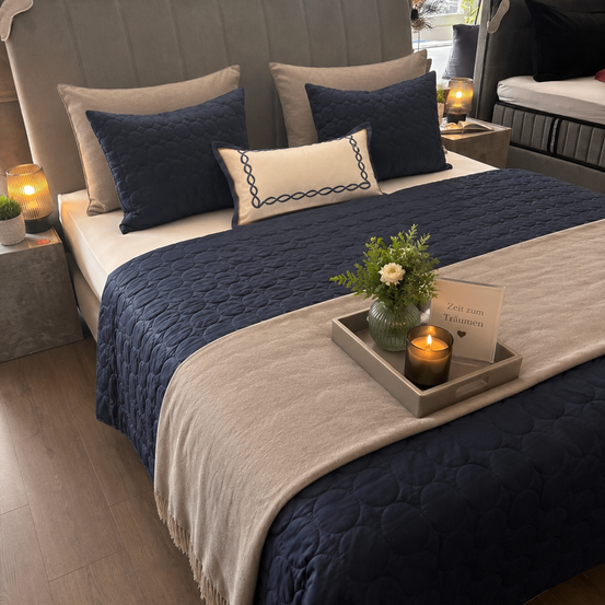 Ein ordentlich gemachtes Bett mit einer blauen Decke, mehreren Kissen und einem Tablett mit einer Kerze und einem Schild, das 'Zeit zum Träumen' auf einem Holzboden steht.