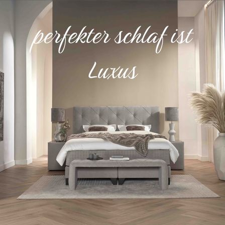 Ein Schlafzimmer mit einem Bett, Lampen und einer Bank davor. Eine weiße Vase mit einer Pflanze und einem Teppich auf dem Boden. Die Wand hat den Text 'perfekter Schlaf ist Luxus.'