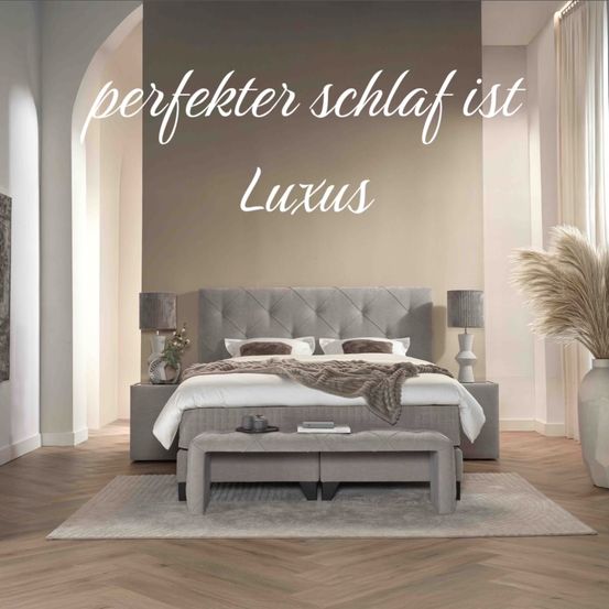 Ein Schlafzimmer mit einem Bett, Lampen und einer Bank davor. Eine weiße Vase mit einer Pflanze und einem Teppich auf dem Boden. Die Wand hat den Text 'perfekter Schlaf ist Luxus.'