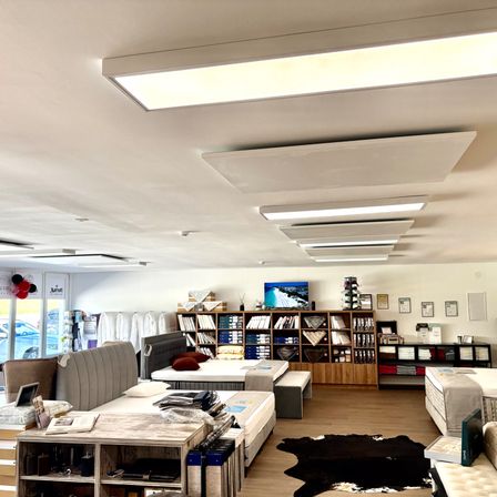 Ein Raum mit mehreren Betten und Möbeln, einem Holzregal mit Büchern und anderen Gegenständen und einem Flachbildfernseher oben. Der Raum hat einen Holzboden und weiße Wände, mit mehreren Deckenlampen und einer Glastür auf der linken Seite.
