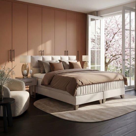 Ein Schlafzimmer mit einem Bett, Kissen und Lampen auf Nachttischen. Es gibt einen Stuhl und einen Tisch mit einer Vase. Der Raum hat einen Holzboden und ein großes Fenster.