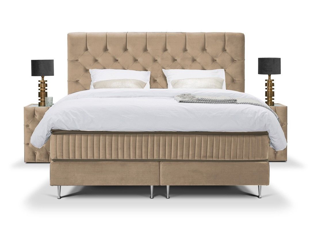 A beige velvet upholstered bed with a quilted headboard, silver metallic accents, and a white duvet. Two nightstands with gold lamps are on each side.