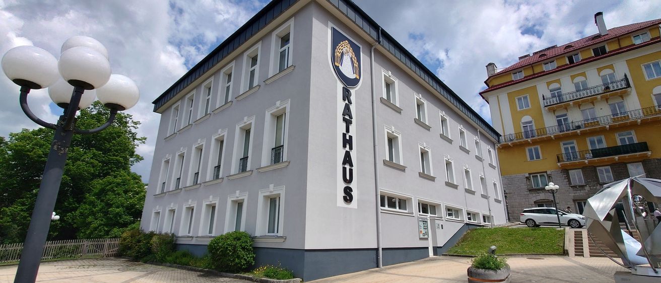 Die Vorderseite eines Gebäudes mit dem Wort Rathaus darauf. Das Gebäude hat viele Fenster und einen grünen Rasen davor.