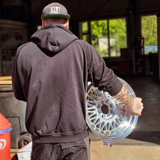 Ein Mann in einer Kappe und einer Kapuzenjacke hält ein glänzendes Alufelgenrad in einer Garage. Das Rad hat eine silberne Felge und gelbe Speichen. Hinter ihm stehen Mülleimer und ein Fenster mit Bäumen draußen.