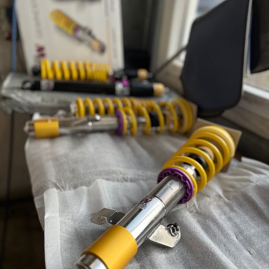 Ein Set gelber und lila Stoßdämpfer wird auf einem Tisch mit einer grauen Stoffabdeckung ausgestellt. Dahinter ist eine Schachtel mit dem Wort 'W' zu sehen.