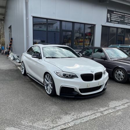 Ein weißer BMW M2 Sportwagen ist vor einem Gebäude mit einer Glaswand geparkt. Ein schwarzes Auto ist daneben geparkt.