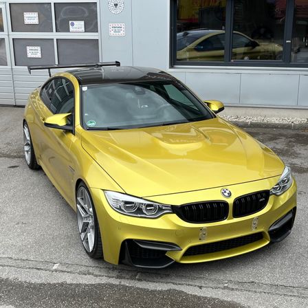Ein gelber BMW M4 Sportwagen steht vor einer Garage mit einer Glaswand, die die Reflexion eines gelben Autos zeigt.