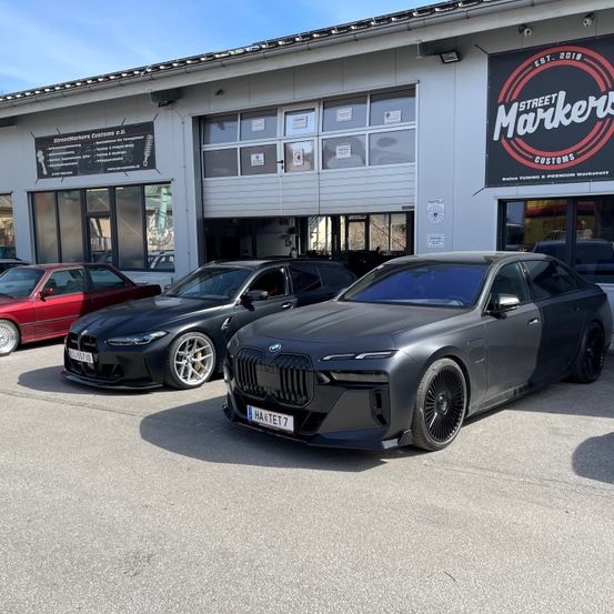 Drei Autos sind vor einem Gebäude mit einem Schild, das 'Street Markers Customs' sagt, geparkt. Die Autos sind alle schwarze BMWs, wobei das vordere Auto das Kennzeichen 'HA ETE 7' hat.