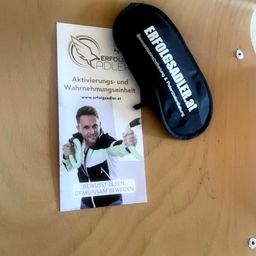 Eine Werbung fur Aktivierungs- und Wahrnehmungseinheit von ErfolgsAdler, mit einem Mann, der eine Fitnessband halt, und einer schwarzen Tasche auf einer Holzoberflache.