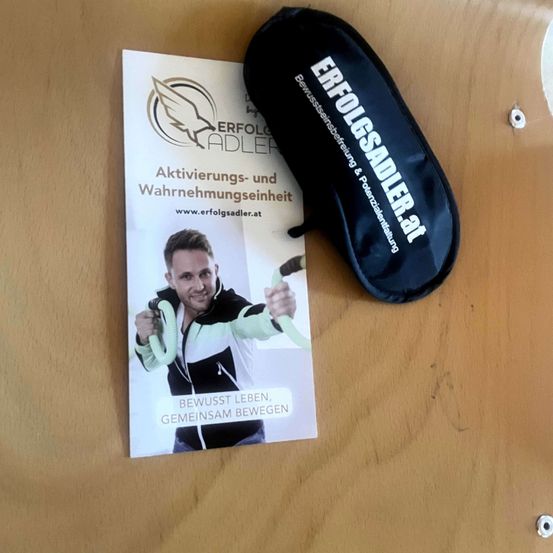 Eine Werbung fur Aktivierungs- und Wahrnehmungseinheit von ErfolgsAdler, mit einem Mann, der eine Fitnessband halt, und einer schwarzen Tasche auf einer Holzoberflache.