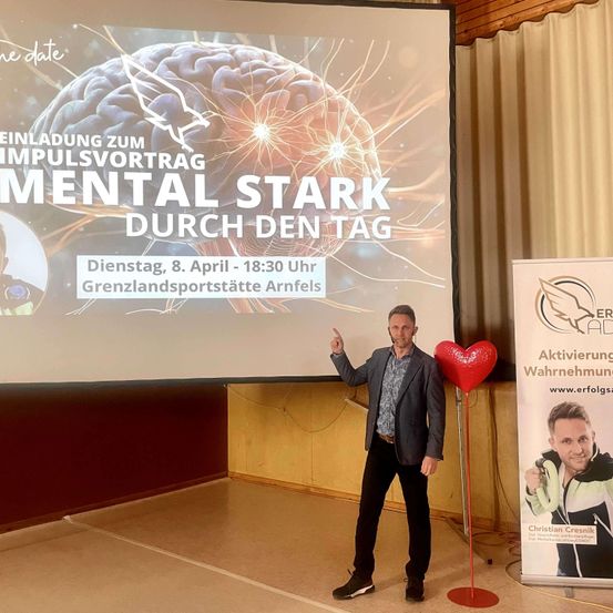 Ein Mann steht neben einem großen roten Herz auf einem Stock vor einer Präsentationsleinwand mit einer Projektion eines Gehirns und zeigt eine Einladung für die Veranstaltung 'Mental Stark Durch Den Tag' am 8. April 2020.