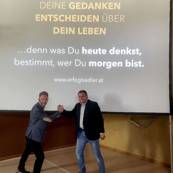 Zwei Männer schütteln sich die Hände vor einer Beamerleinwand mit einer motivierenden Botschaft.