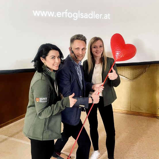 Drei Menschen stehen zusammen, lächeln und posieren für ein Foto mit einem roten Herz. Ein Mann hält das Herz, und eine Frau steht links. Hinter ihnen ist eine weiße Wand mit einer Website-Adresse.