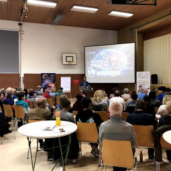 Ein Raum mit einer Projektionsleinwand, einem Mann am Rednerpult und einer Menschenmenge in Stühlen sitzend.