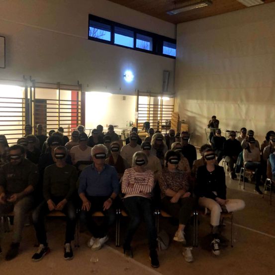 Eine Gruppe von Menschen sitzt in Stühlen mit verbundenen Augen und beobachtet eine Präsentation in einem Raum mit weißer Wand und Holzdecke.