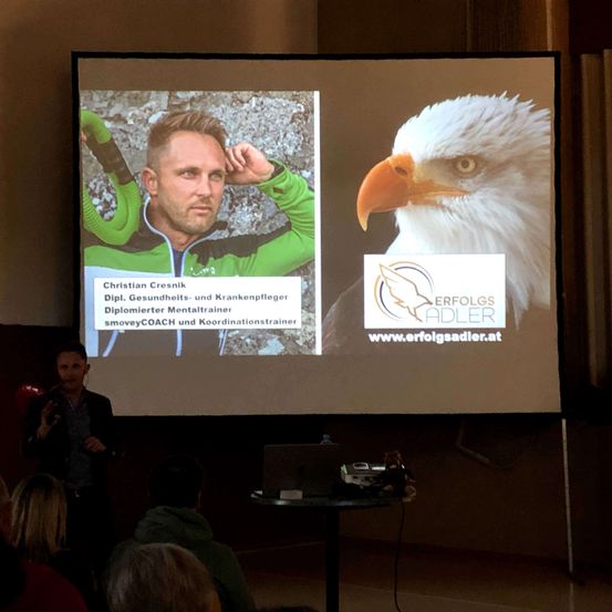 Ein Mann hält eine Präsentation, hinter ihm ist eine Leinwand mit einem Bild eines Mannes in einem grünen Jackett und einem Adler.