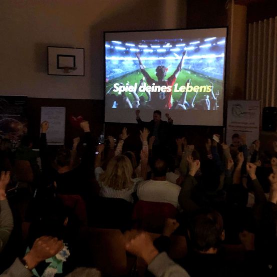 Publikumsmitglieder in einem großen Raum mit einer Projektionsleinwand, die einen Fußballspieler mit erhobenen Armen und den Worten 'Spiel deines Lebens' zeigt.