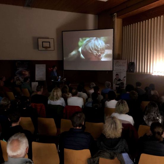 Eine große Gruppe von Menschen sitzt auf Stühlen und schaut sich eine Präsentation auf einer großen Leinwand an. Eine Person steht auf der Bühne und präsentiert wahrscheinlich etwas. Ein Basketballkorb ist an der Wand hinter der Person montiert.