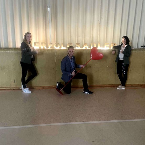 Drei Personen posieren vor einer Wand mit einem Schild, das 'Erfolgssalder' steht, wobei eine Person auf einem Knie steht und ein großes rotes Herz hält.