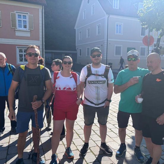 Eine Gruppe von Wanderern, gekleidet in Outdoor-Bekleidung, darunter Hüte, Sonnenbrillen und Rucksäcke, posiert für ein Foto auf einer Kopfsteinpflasterstraße. Ein Baum und ein Gebäude mit Fenstern sind im Hintergrund zu sehen.