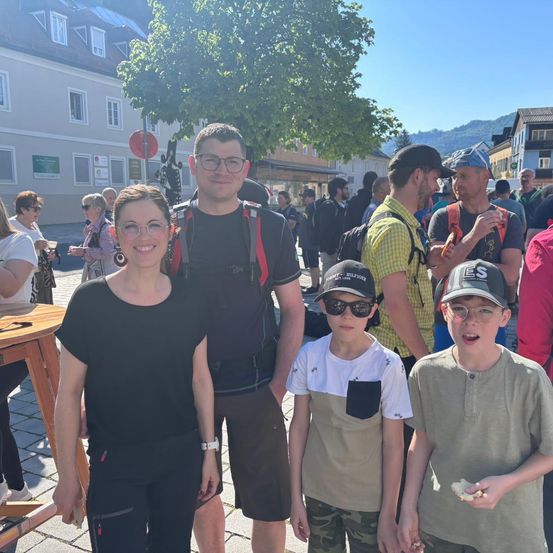 Eine vierköpfige Familie steht vor einer Menschenmenge in einer Außenumgebung. Der Mann trägt einen Rucksack und eine Brille, und die Frau trägt eine Uhr. Die beiden Jungen tragen Mützen und Hemden. Im Hintergrund befinden sich Gebäude und Bäume.