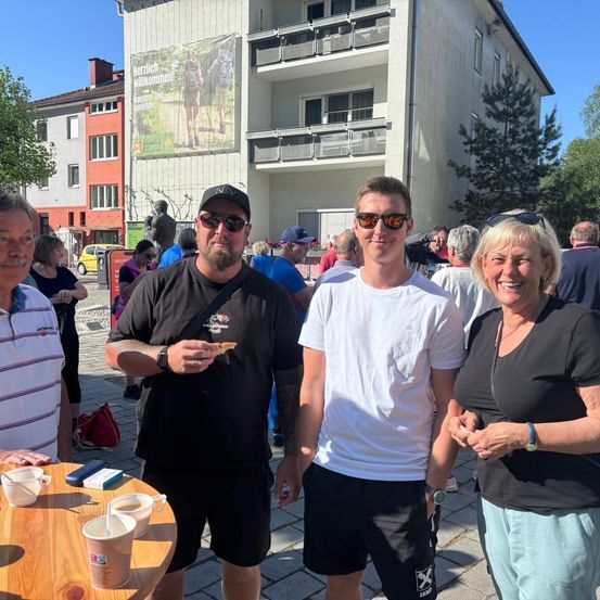 Drei Menschen stehen vor einem Tisch mit Tassen und einem Mann, der ein Stück Essen in der Hand hält. Sie lächeln und scheinen für ein Foto zu posieren. Dahinter befinden sich Gebäude, Bäume und andere Menschen.