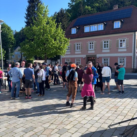 Eine Gruppe von Menschen versammelt sich vor einem Gebäude mit Solarpanelen auf dem Dach. Einige tragen Hüte, Turnschuhe und Rucksäcke.