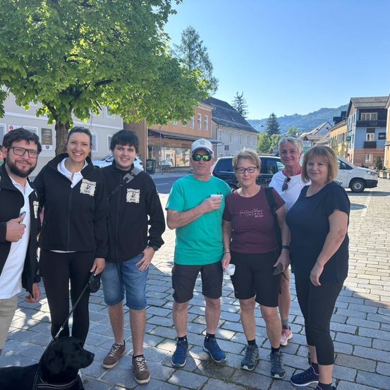 Eine Gruppe von Menschen steht auf einer Kopfsteinpflasterstraße und lächelt, ein Hund neben ihnen. Sie sind in einer Stadt mit Gebäuden und Bäumen im Hintergrund. Ein Mann hält einen Becher, und andere haben Rucksäcke. Ein Van ist in der Ferne geparkt.