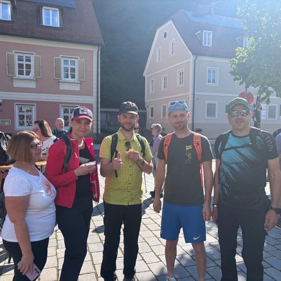 Fünf Menschen stehen vor einem Gebäude mit gepflastertem Boden, einer von ihnen gibt einen Daumen hoch, und sie tragen alle Hüte und Brillen.