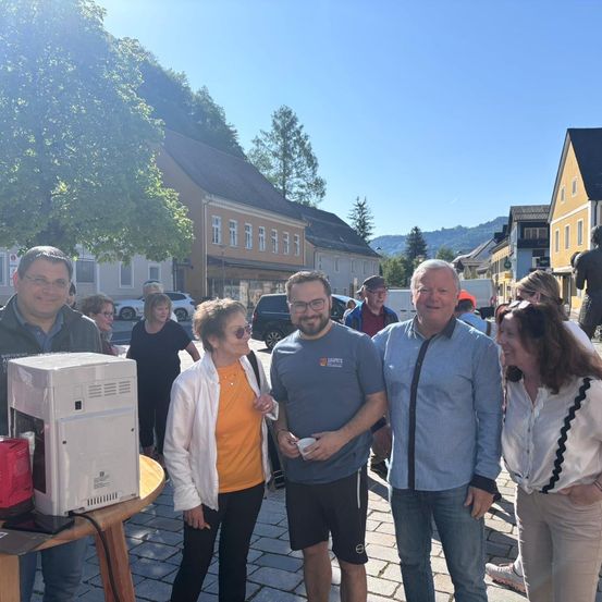 Fünf Personen stehen auf einem kleinen Platz mit gepflastertem Boden. Sie lächeln und unterhalten sich. Ein Mann hält einen Becher. Auf einem Tisch steht eine Kaffeemaschine. Im Hintergrund gehen Menschen vorbei.