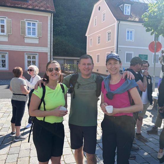 Drei Personen posieren für ein Foto auf einem Stadtplatz. Die Frau auf der linken Seite trägt ein neongrünes Shirt, und der Mann in der Mitte trägt ein grünes Shirt. Die Frau auf der rechten Seite trägt ein rosa Shirt. Sie alle halten weiße Tassen. Hinter ihnen sind weitere Personen und Gebäude.