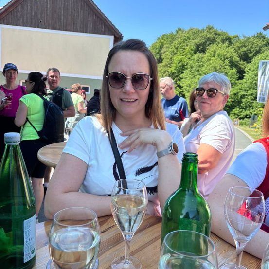 Eine lächelnde Frau sitzt an einem Tisch mit zwei Weingläsern, einer Flasche Wein und einer weiteren Flasche. Sie trägt eine Sonnenbrille und eine Uhr. Hinter ihr stehen und sitzen andere Menschen in einem Außenbereich.