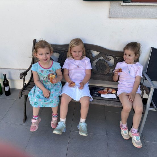 Drei junge Mädchen sitzen auf einer Holzbank, lächeln und halten Essen. Sie sind in bunte Kleidung gekleidet. Die Bank steht auf einem gefliesten Boden.